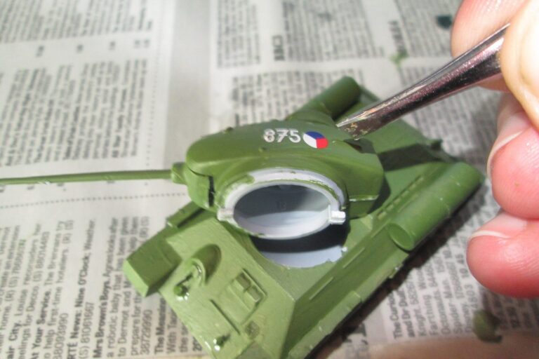 Modellbauer bringt mit einer Pinzette ein Decal auf ein grünes Panzermodell auf