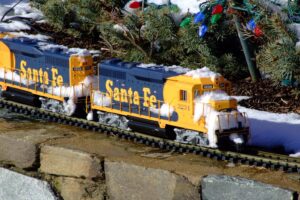 Gelbe Modelllokomotive auf verschneiter Gartenbahn unter einem beleuchteten Weihnachtsbaum